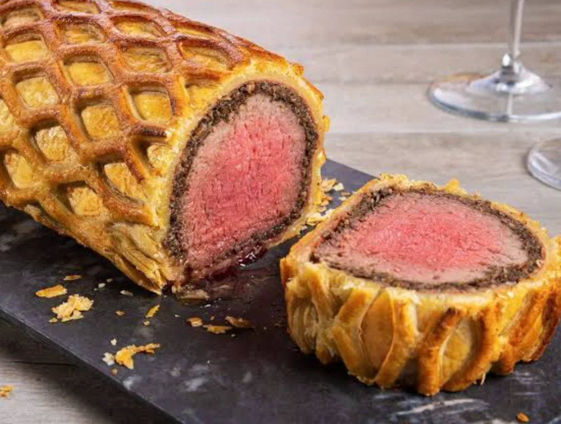 A beef Wellington in latticed puff pastry on a slate board, cut to show the pink beef fillet and dark duxelles inside.