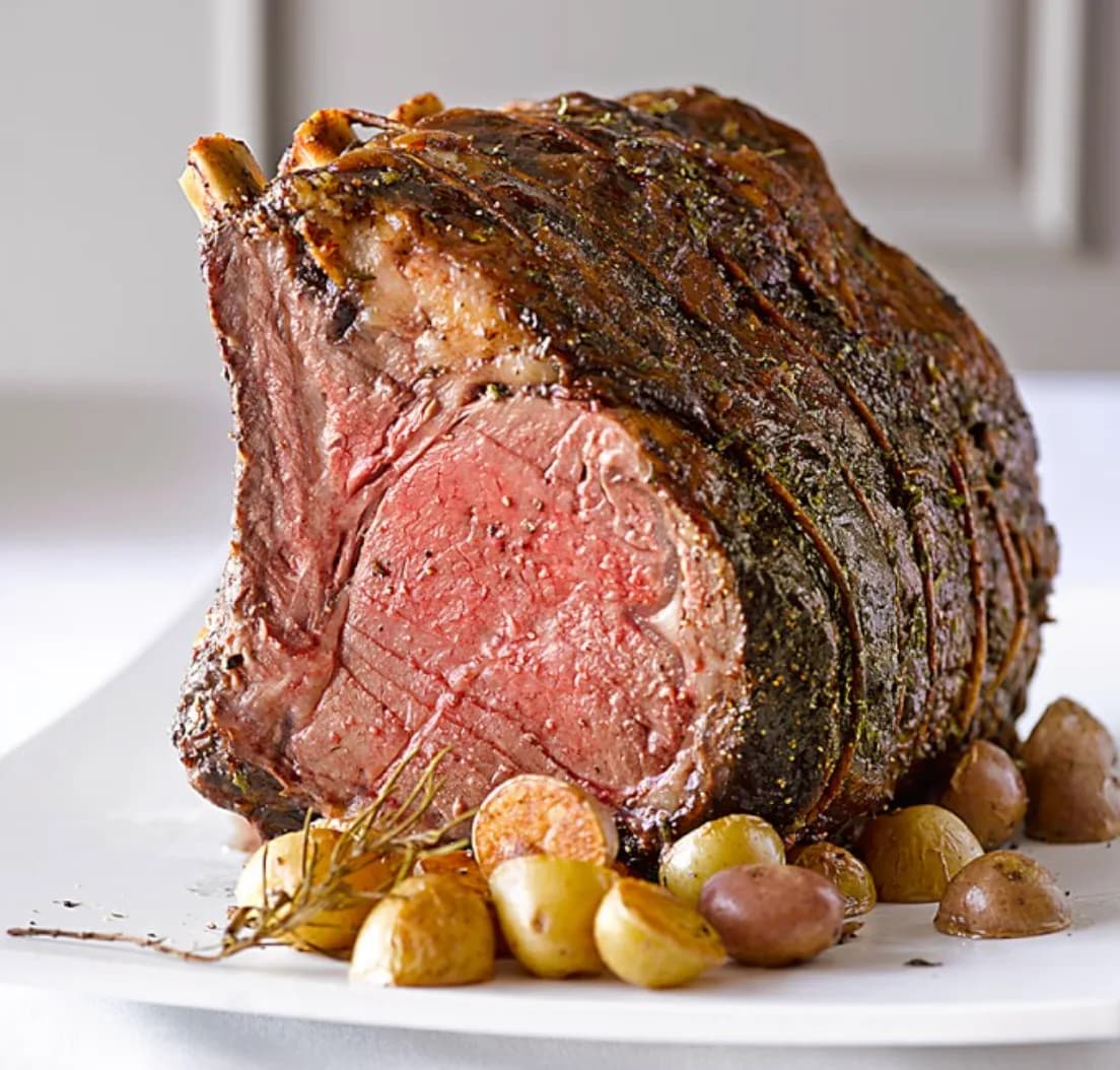 A standing bone-in rib roast with a herb crust, the cut end showing pink medium-rare meat, served on a white platter with roasted baby potatoes.