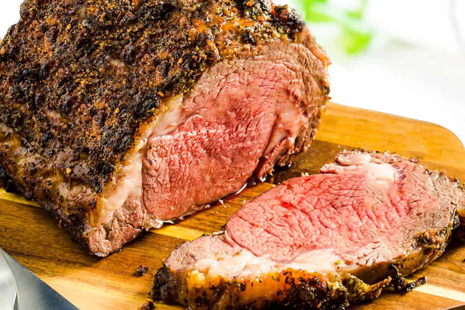 A boneless rib roast with a dark herb and pepper crust on a wooden board, two thick slices carved from the end showing pink meat.