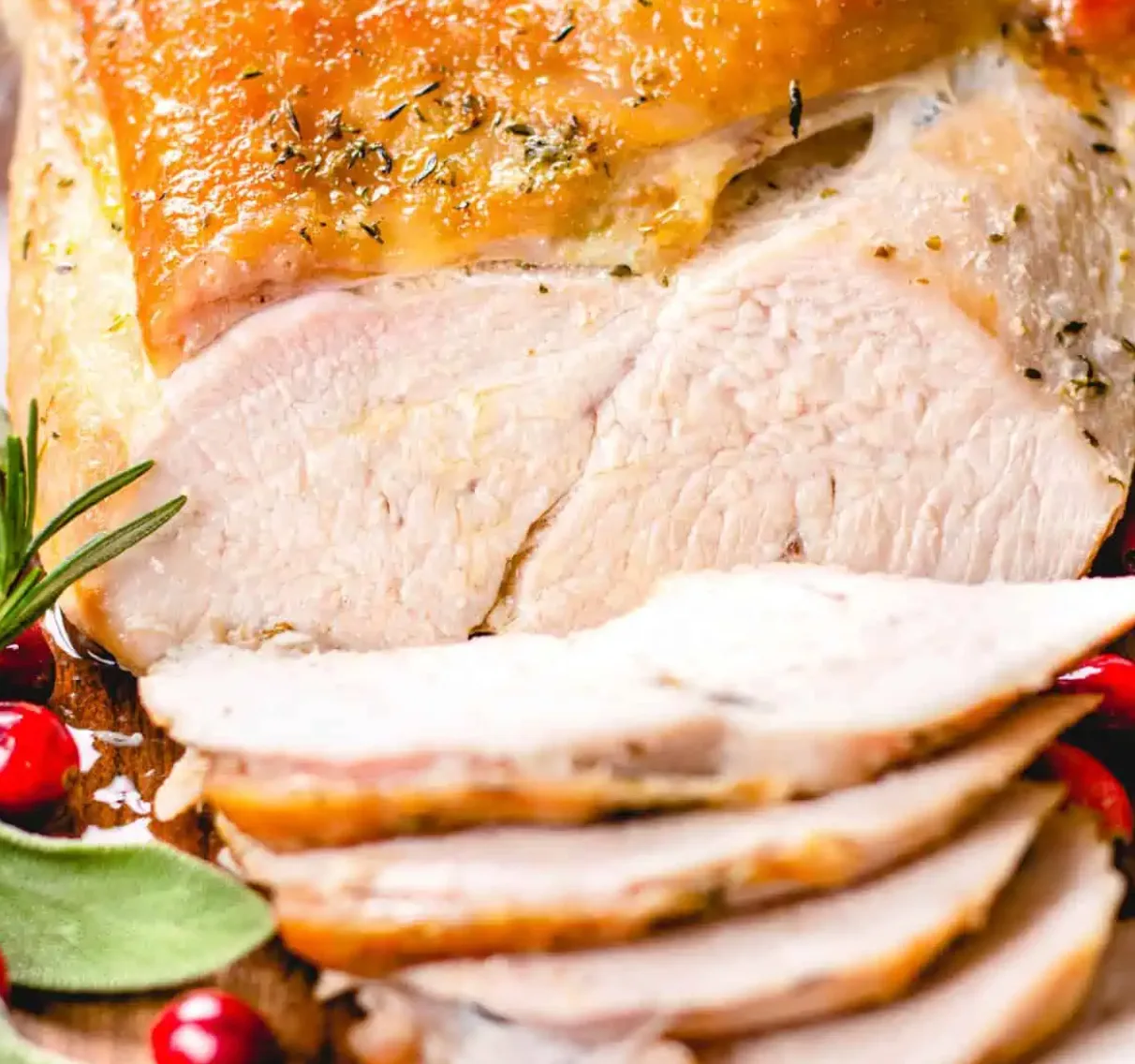 A roasted turkey breast with golden crisp skin, carved into thick slices and garnished with rosemary, sage and cranberries.