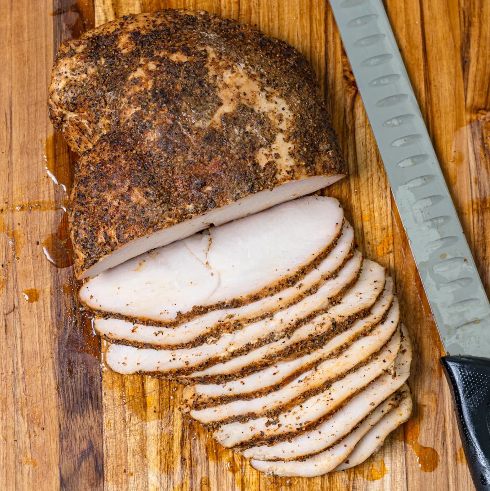 A smoked turkey breast with a dark peppery crust on a wooden board, part of it carved into thin slices beside a carving knife.