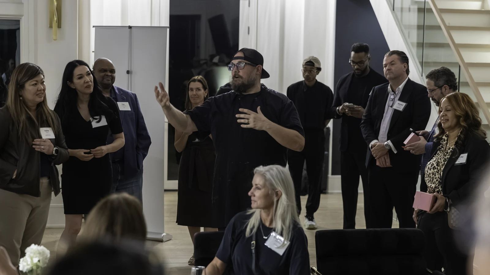 Chef Mark Frazie talking to a room of guests wearing name badges at a corporate event, mid-sentence with one hand raised.