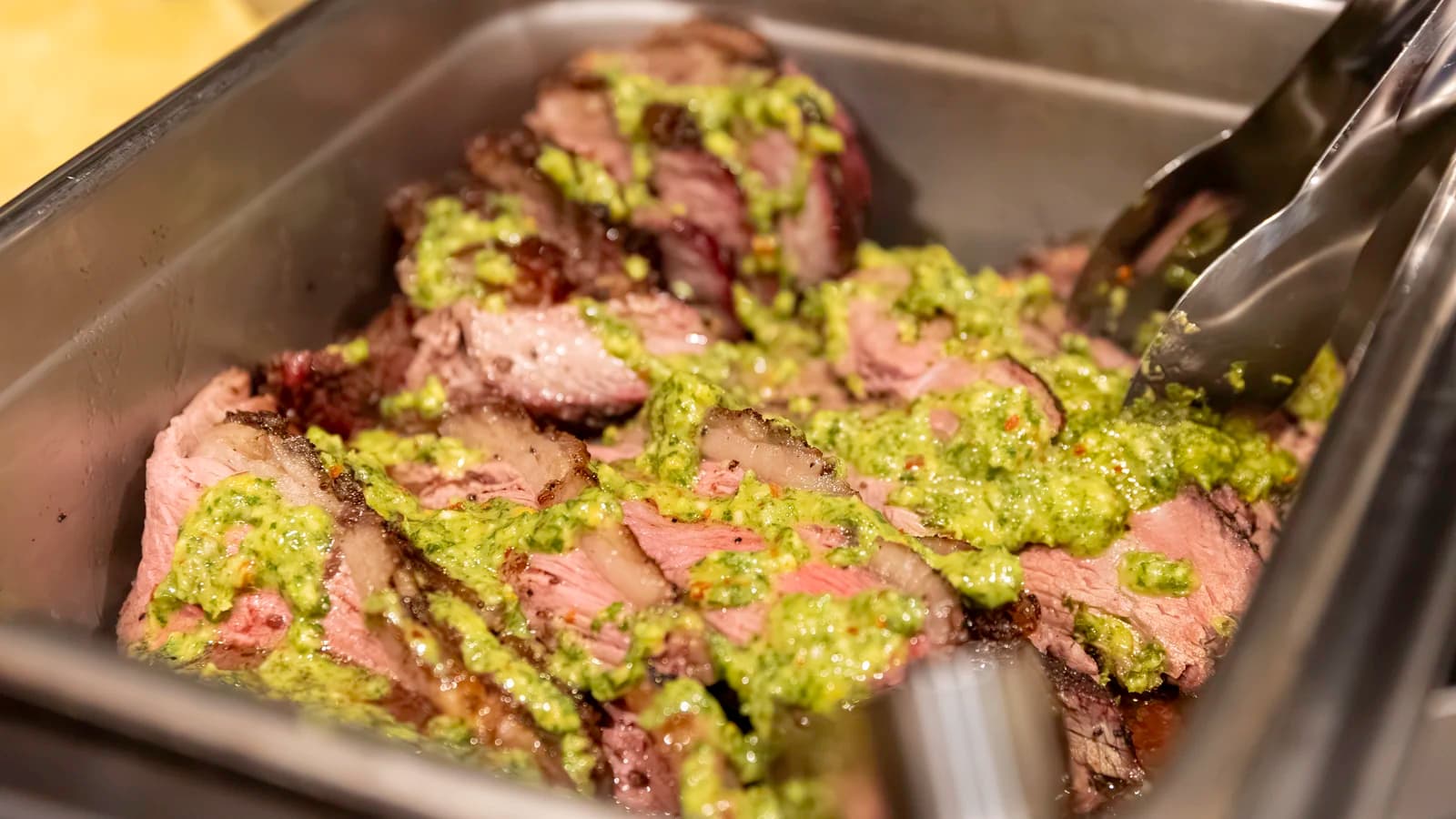 Sliced beef with a peppered crust under green chimichurri sauce, held warm in a chafing dish with serving tongs.