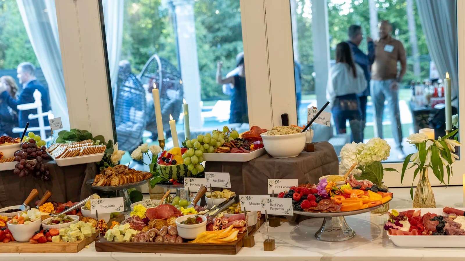 A long grazing table of charcuterie, cheeses, fruit and dips on wooden boards, laid out beside tall windows with guests outside.