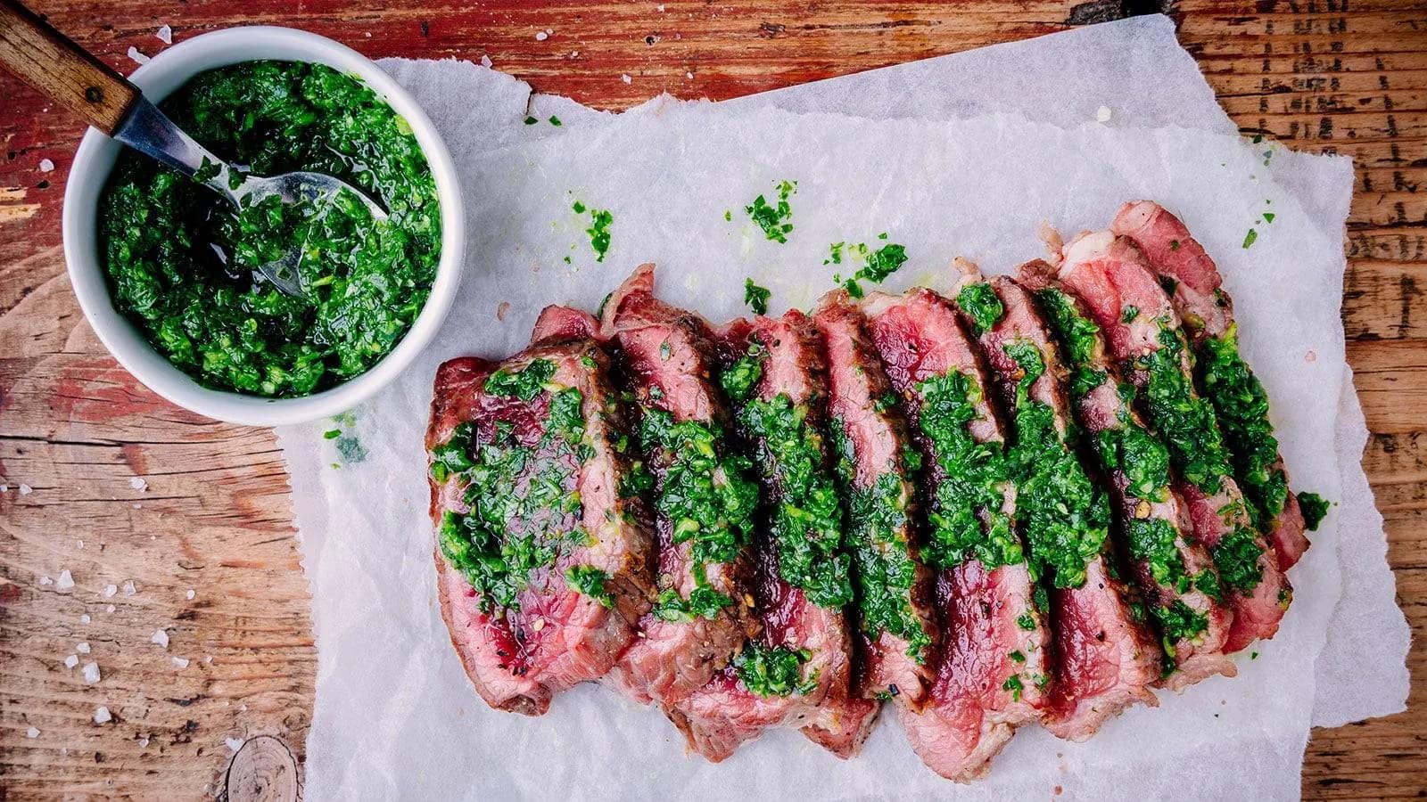 Slices of chimichurri encrusted beef tenderloin laid out on parchment paper with green chimichurri sauce spooned over them.