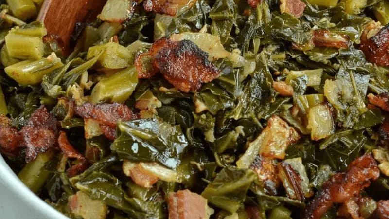 BBQ braised collard greens with pieces of bacon in a dark, rich braising liquid.