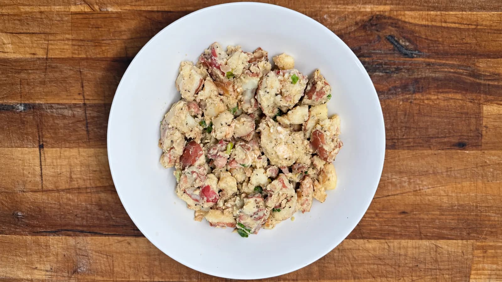 German potato salad with red skinned potato chunks, bacon and sliced scallions in a creamy dressing, served in a white bowl on a wooden surface.