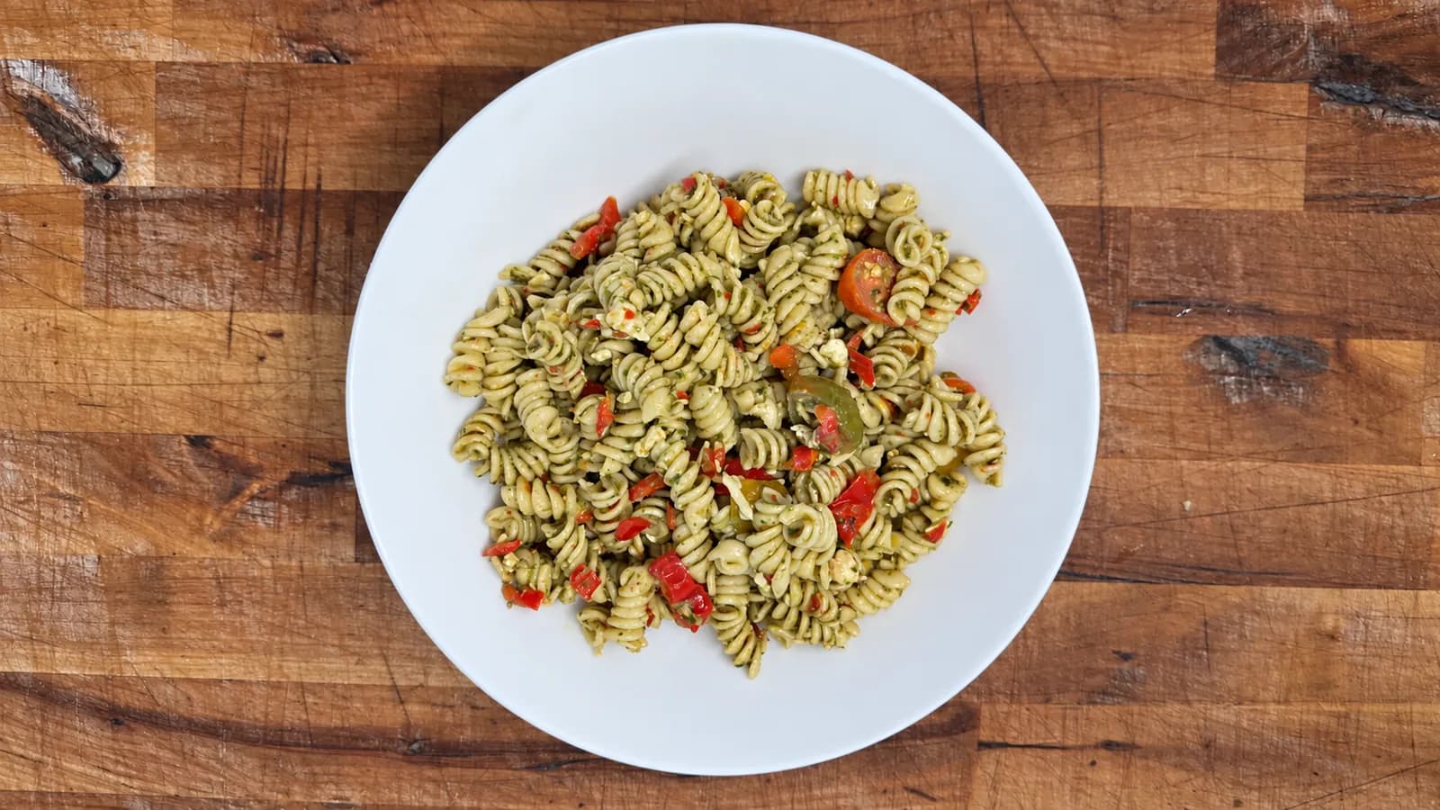 Pasta salad with spiral rotini, roasted red peppers, green olives and herbs, served in a white bowl on a wooden surface.