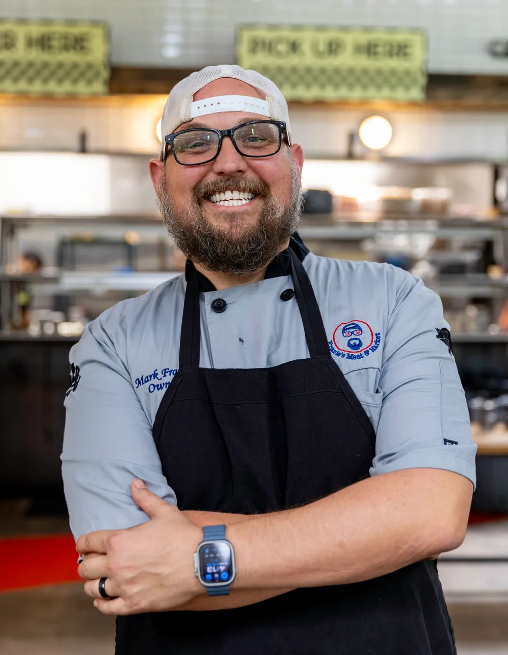 Mark Frazie, owner of Frazie's Meat & Market, in a chef's jacket and apron with his arms crossed, at the counter in the shop.