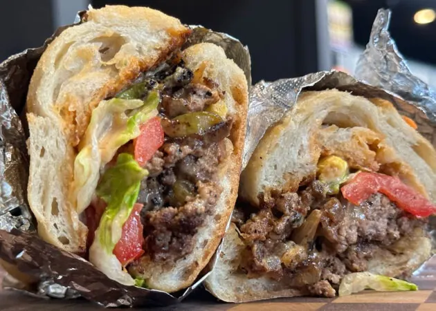Gourmet sandwich featuring ground beef, fresh lettuce, tomatoes, and pickles, wrapped in foil, highlighting Frazie's Meat & Market's hand-crafted offerings.