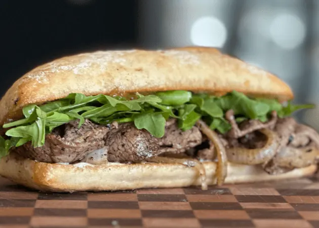 Hand-crafted sandwich featuring tender beef, sautéed onions, and fresh arugula on artisan bread, representing Frazie's Meat & Market's gourmet offerings in Atlanta.