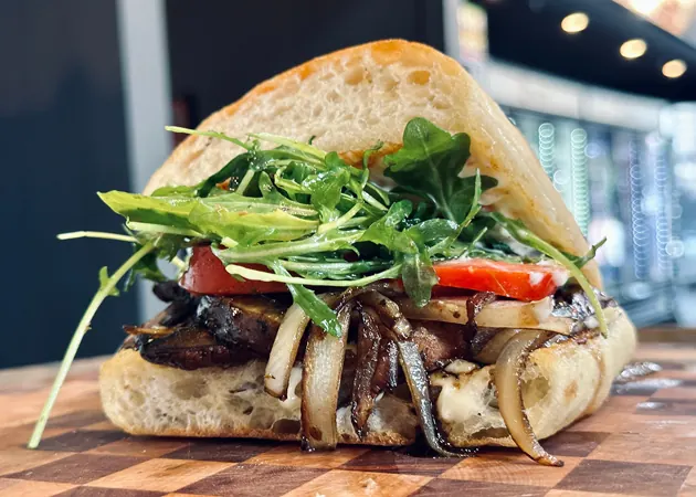 Hand-crafted gourmet sandwich featuring grilled mushrooms, onions, fresh arugula, and tomato on rustic bread, highlighting Frazie's Meat & Market's commitment to high-quality, locally sourced ingredients.