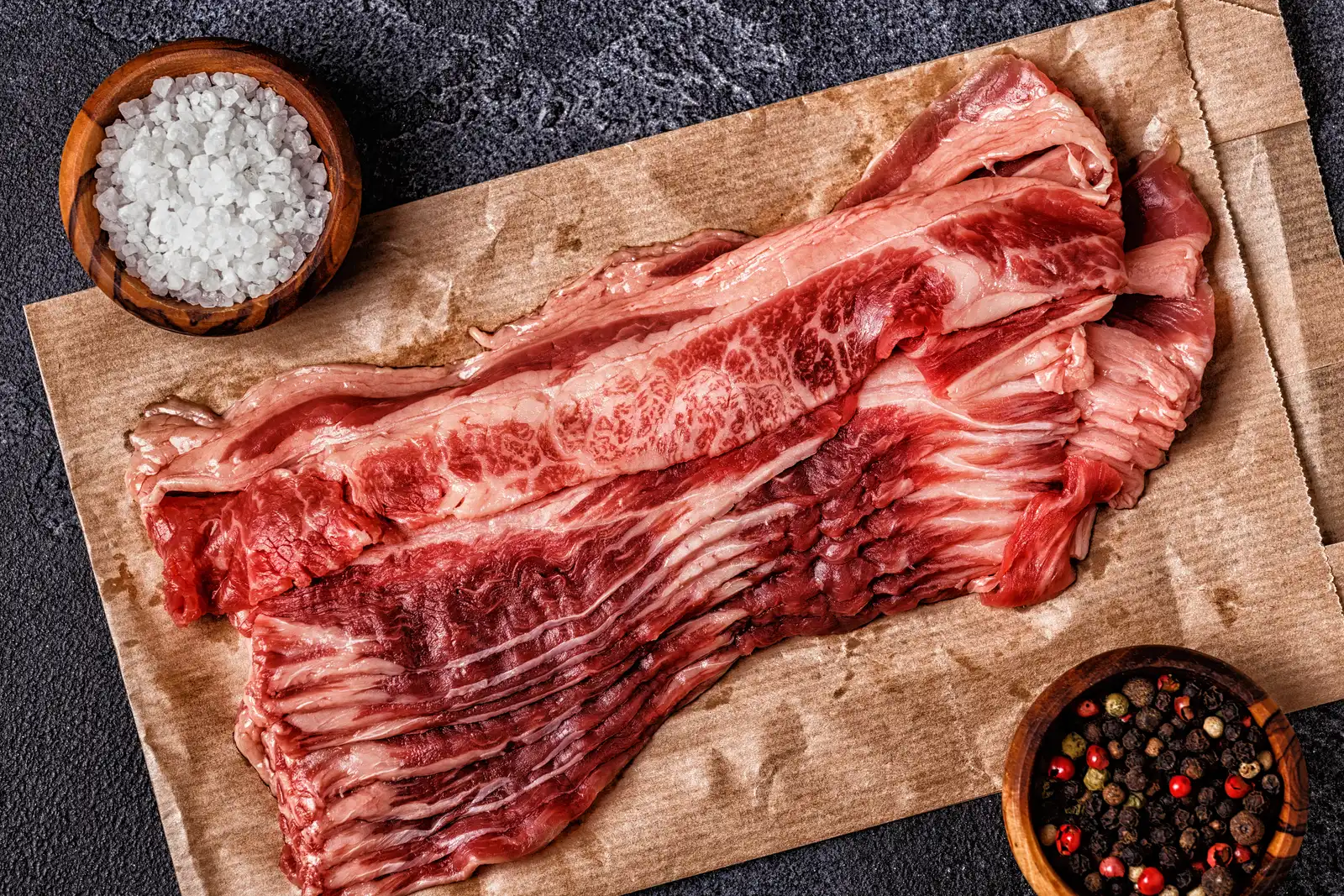A slab of raw beef bacon on butcher paper beside bowls of coarse salt and whole peppercorns, ready for the cure.
